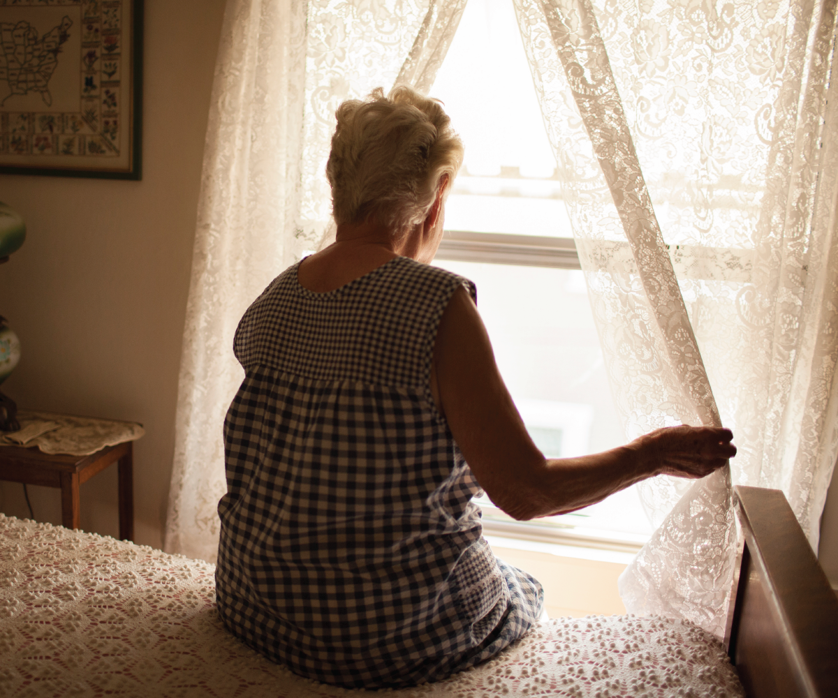 Elderly hispanic woman sits on her bed and looks out the window.
